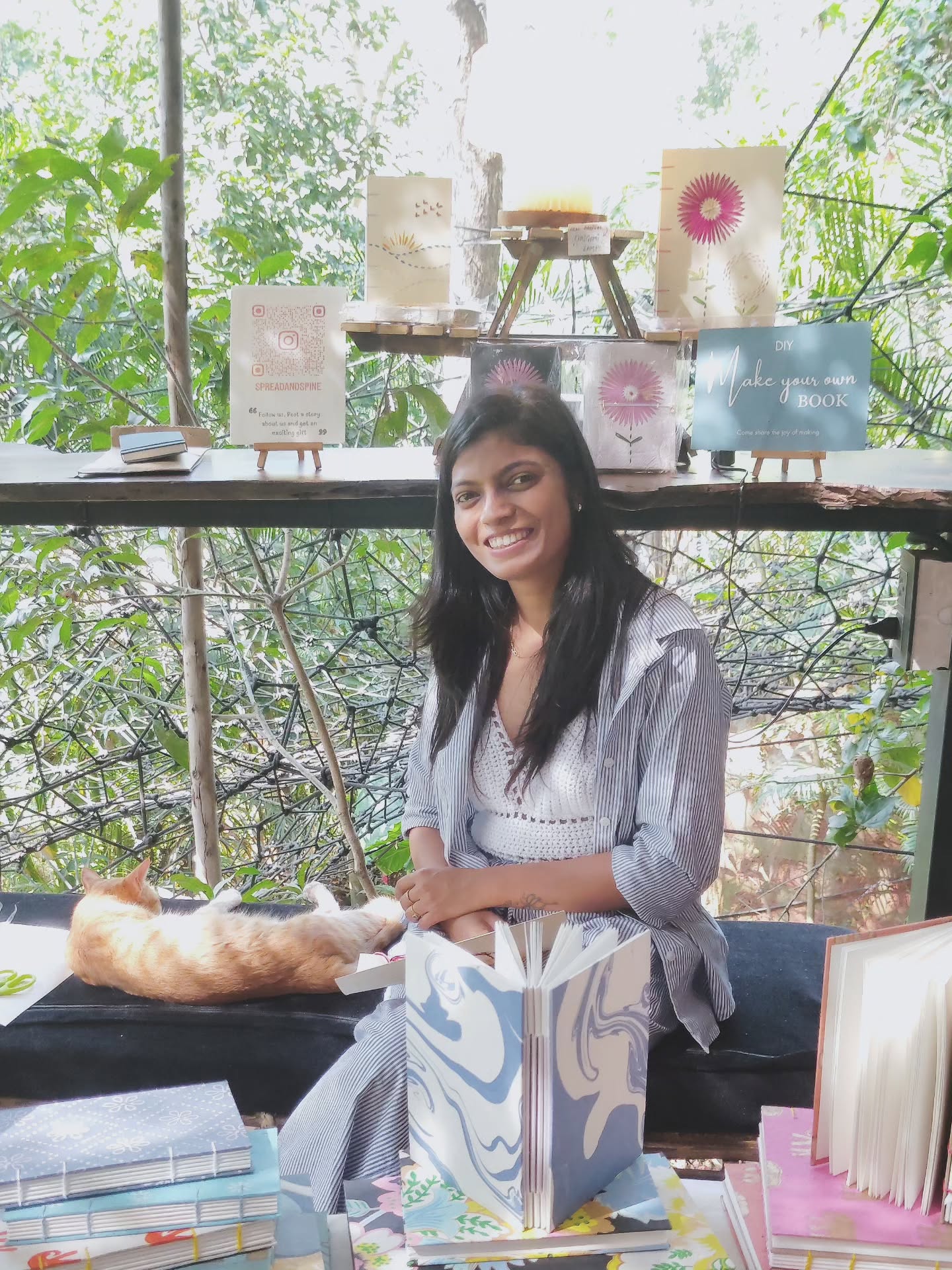 The founder of Spread & Spine smiling at her studio stall, stitching a marbled Coptic journal with a ginger cat beside her.